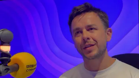 Rob Spring sits in a radio studio beside a yellow BBC‑branded microphone. he is front a blue and purple patterned backdrop. He has short brown hair and stubble