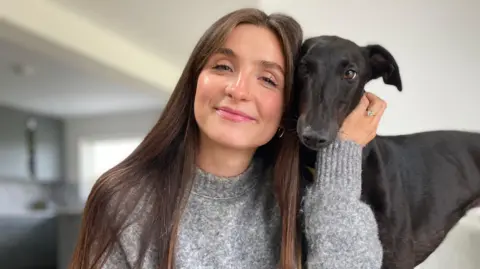 A young woman who is smiling, she has brown hair and is wearing a grey jumper. A black greyhound is standing beside her and is leaning slightly against the side of her head. 