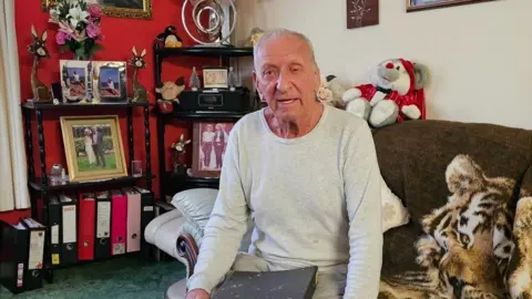 An older man wears a light grey long-sleeved top and sits on a sofa holding a dark file or binder. Behind him are shelves containing framed family photographs, flowers, ornaments and ring binders. A patterned throw covers part of the sofa and soft furnishings fill the room.