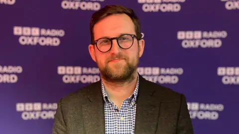 Freddie van Mierlo stands in front of a purple banner with BBC Radio Oxford branding. He has short dark hair, black rimmed glasses, and wears a dark suit jacket with a checked shirt.