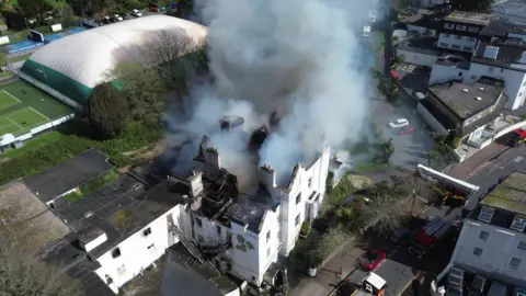 An ariel view of smoke bellowing from the derelict hotel. The building is white and the roof is black with smoke.