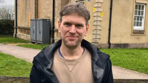 A middle-aged man with dark grey hair and stubble is standing outside of a building smiling. He is wearing a black raincoat and beige sweatshirt.