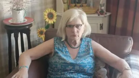 Janice Lees A woman in a blue patterened v-neck dress with bead necklace and turquoise bracelet. She is sitting in a leather chair in a room with checked wallpaper and a tall sideboard behind her with fake sunflowers and ornaments.