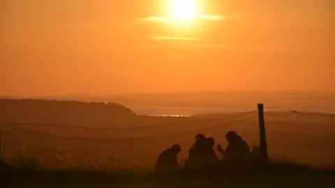 Mendip Basecamp Three people are silhouetted against an orange sky during sunset. They are sitting on a hill overlooking a valley.