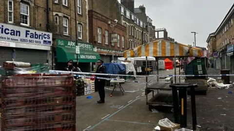 Police tape surrounds a market area, with debris scattered and officers investigating.