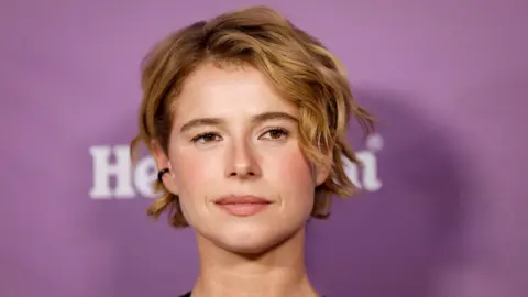 REUTERS/Caroline Brehman Un primo piano di Jessie Buckley che partecipa al BAFTA Tea Party 2026 a Los Angeles. Ha i capelli biondi corti e ondulati ed è raffigurata davanti a uno sfondo viola. 