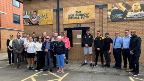 A group of people stand either side of a silver plaque outside the Rob Burrow Fitness Suite.