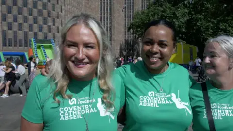 A blonde woman smiling in front of a brown brick cathedral. She is wearing a green t-shirt with white writing that reads "Coventry cathedral abseil".
