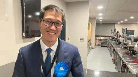Amy Holmes/BBC A man in a smart blue suit and tie is standing indoors, smiling toward the camera. A blue rosette pinned to the jacket clearly reads “Conservatives.” The setting appears to be a bar or club area, with multiple drink taps, a counter, and glassware visible in the background.