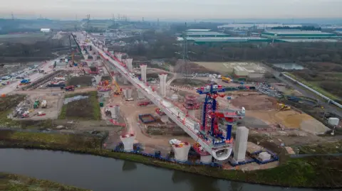 HS2 A large grey viaduct covered in cranes, red cones and construction traffic and equipment. It runs towards a river. There are pylons nearby and large industrial buildings in the background.