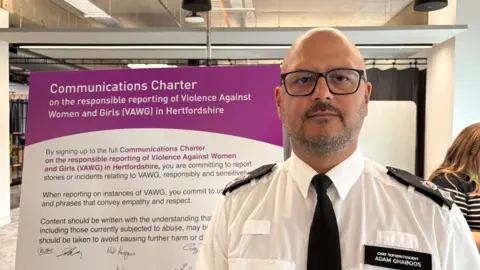 Martin Heath/BBC Adam Ghaboos with very short dark hair and slight beard. He is wearing black framed glasses and is looking at the camera with a neutral expression. He is wearing a white police shirt with black epaulettes and a black name patch on the left pocket. He is standing in front of a noticeboard setting out a communications charter.