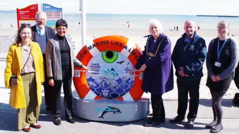 A group of six people standing either side of an installation shaped like a lifebuoy as they unveil the new unit