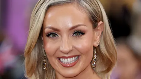 Close up of smiling blonde women with sparkly, dangly earrings against a blurred background.