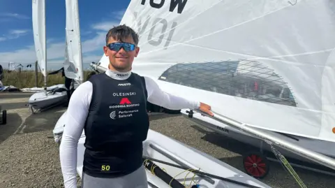 A smiling Arthur wearing light blue reflective sunglasses, a black waterproof vest with a white long sleeved wetsuit. He has placed his left arm on a boat.