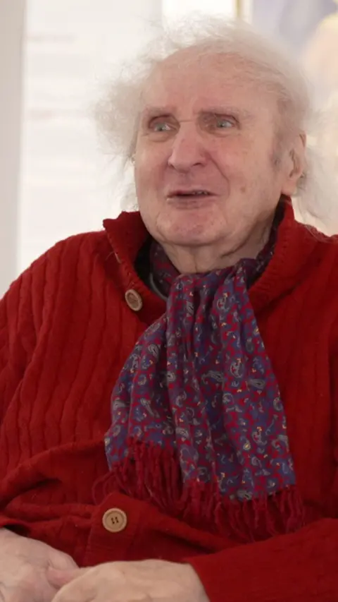 A man with white hair is wearing a red cardigan and a floral scarf.