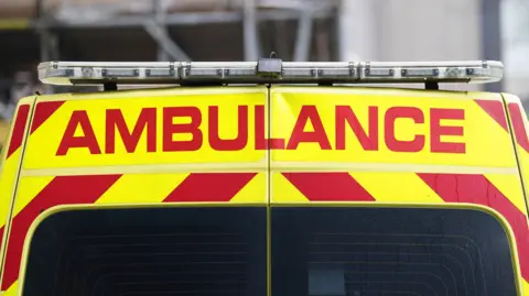 The back of a yellow and red ambulance parked in front of a blurred building which has scaffolding outside of it. The word "ambulance" is written is red block capitals.