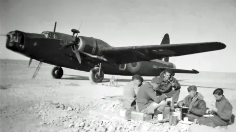 Phil Le Brocq A black and white photograph showing a group of five uniformed men sitting on the floor of a bar eating. Behind them is a Wellington bomber.