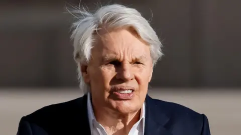 Reuters Mike Jeffries, an elderly man with white hair, wearing a dark suit and a white -open-necked shirt, photographed with a long lens outside the federal courthouse in Central Islip, New York, where he was arraigned on charges of sex trafficking and prostitution