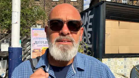 BBC/ Seb Cheer An older man with a bald head, white beard, and dark sunglasses stands outdoors in bright sunlight, wearing a blue checkered shirt and holding a shoulder strap, with stone buildings and posters behind him.