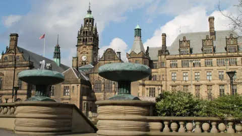 Sheffield town hall, a sandstone-coloured building that stretches across several floors. On the left, it features a clock tower which features a bronze statue on the roof's spire. 