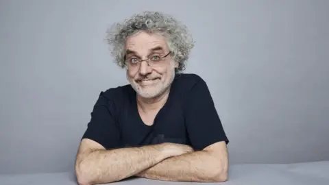 Aardman Animations Studio picture of Matt Perry - he has curly grey hair and glasses, smiling with arms crossed in front of him. He is wearing a blue t-shirt and has a small hoop earring in his left ear.