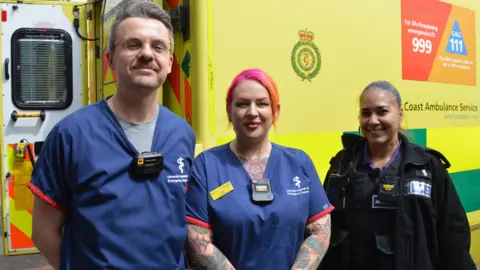 University Hospitals Sussex NHS Trust Three NHS staff members, two wearing hospital scrubs and a third wearing ambulance trust uniform with all three wearing body-worn cameras on their uniform while standing in front of an ambulance.