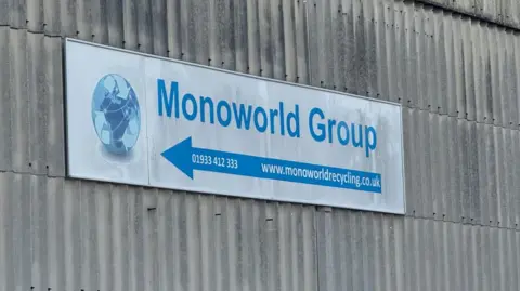 Steve Hubbard/BBC Monoworld sign, with Monoworld Group in blue lettering with a logo, phone number and website address. The sign is attached to a corrugated iron warehouse wall.