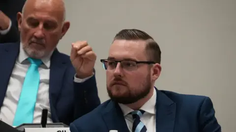 A bearded man is wearing a white shirt, with a striped tie and a light navy blazer. His hair is gelled to the right side of his head in a side parting. He is thoughtfully looking across the council chamber
There is another man behind him also wearing a suit and tie