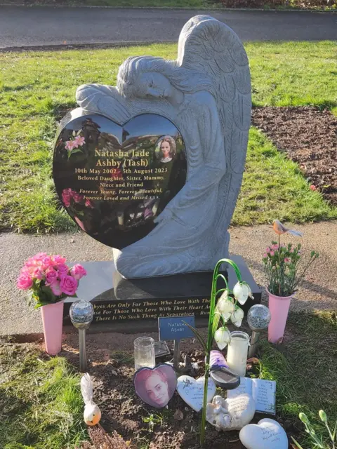 Family The image shows a grave marked with a carved stone angel holding a heart. Flowers and pictures surround it