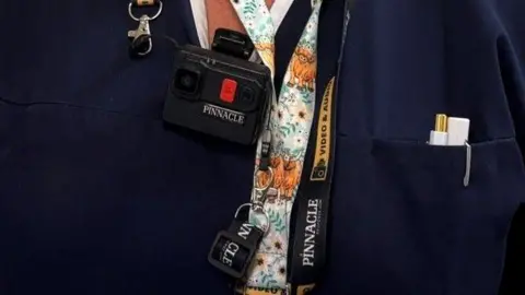 An NHS worker has a lanyard on reading 'Pinnacle' and a black body camera on. They have two white pens in their uniform pocket.