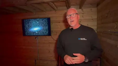 Gary Fildes, director of Grizedale Observatory, standing next to a monitor displaying a picture of the Andromeda galaxy. It is a bright spiral in the middle of the screen and is surrounded by stars.