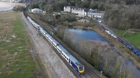 Looking down across a train which has derailed on its way to Barrow-in-Furness, in Cumbria. The train is Northern operated, with blue and yellow branding. 