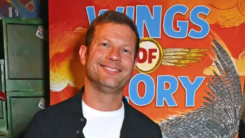 Getty Images A man with short brown hair, a white t-shirt and navy jacket in front of a large orange poster with the words 'Wings of Ory' on it