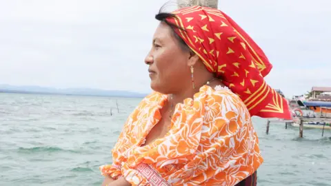 Side view of Yanisela, wearing a red and yellow headscarf and an orange and white floral blouse. She is looking out to sea with some jetties and buildings on the island behind her.