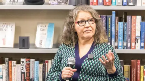 Angela Montgomery holding a microphone and speaking in front of bookshelves filled with various books in a bookstore setting.