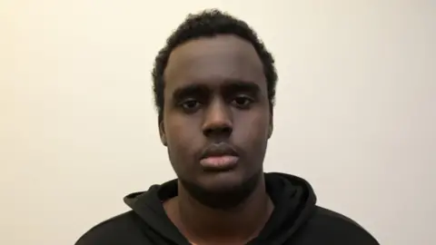A custody mugshot of Mohamed Mohamoud, who has a serious expression on his face as he stares at the camera. He has short curly black hair and is wearing a black hoodie