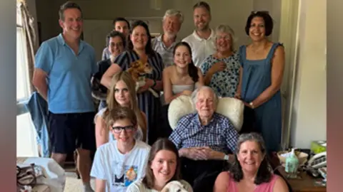 Family handout A large family group, in a house, with a man sitting in a chair, all round him. A person at the front is holding a dog, as well as a woman to the left. They are all smiling and looking at the camera.