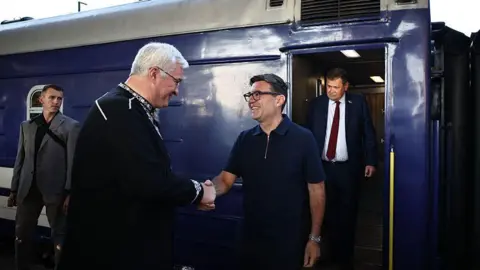 Burnham shakes the hand of Sybiha as they both smile and look forward while stood next to two other men and a train