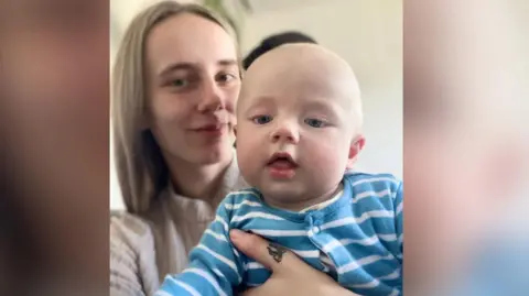 Lauren Parrish holds her son Archie up to the camera. Archie is wearing a blue striped top