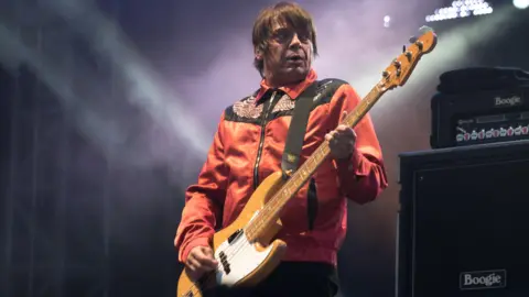 Getty Images Mani playing bass on stage, wearing a red jacket