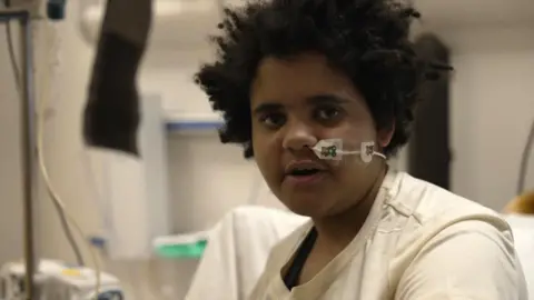 Grace, sat on her bed, with medical equipment blurred in the background. 