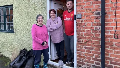 Naj Modak/BBC Sylvia Fisher, a woman in a pink jumper and blue trousers and wellies, with a couple at the door of a terraced house which has flooded 