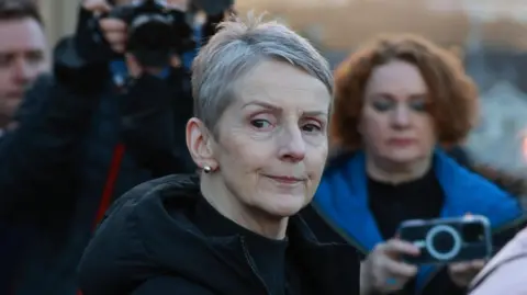 A close-up image of Lady Eleanor Donaldson outside court in Newry. Behind her are reporters with cameras and phones pointed at her. Lady Donaldson is an older woman with short grey hair. She has a neutral expression on her face. She is wearing a large, black coat.