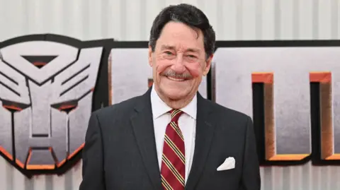 Peter Cullen standing and smiling in front of a Transformers logo backdrop