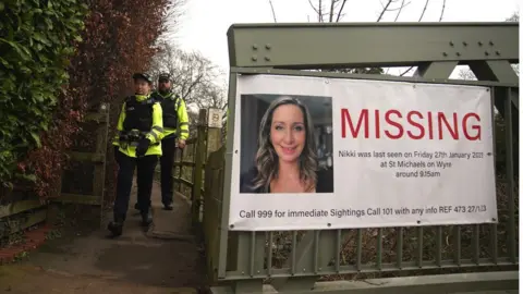 PA Media Police officers walk by a poster appealing for information about the disappearance of Nicola Bulley, on a bridge over the River Wyre