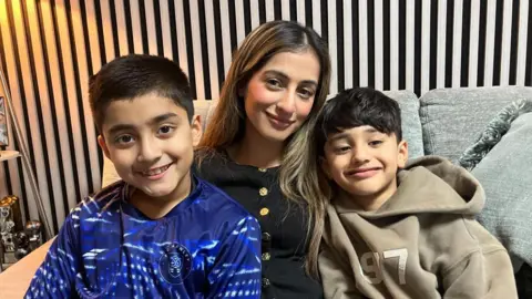 Two boys with short dark hair, one in a blue top and one in a beige top, sitting on either side of a woman with long brown hair in a black top