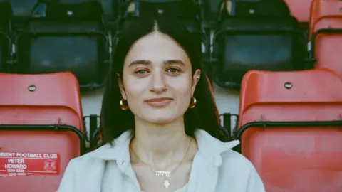 Natine Williams Marva is sat in the stands at a football stadium with red and black seats in the background. She is wearing a white top shirt with a white vest top underneath and gold earrings, staring straight at the camera
