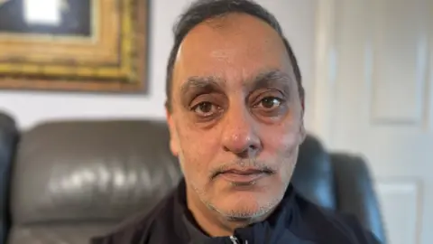 BBC Araf Saddiq stares at the camera with a serious, sad expression, He is wearing a black jacket zipped up to the neck. He is sitting on a leather couch in a domestic setting with a gold framed painting visible in the background.
