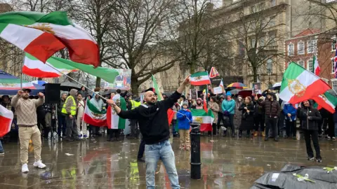 A man is waving the Iranian flag in front of a crowd of people also waving Iranian flags. They are standing in a square which is wet from rain.