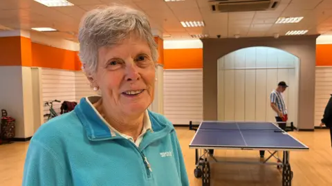 Valerie Lawrence is smiling at the camera and standing in an open space with a table tennis table in the background and a wooden floor. She has short, grey hair and is wearing a blue top
